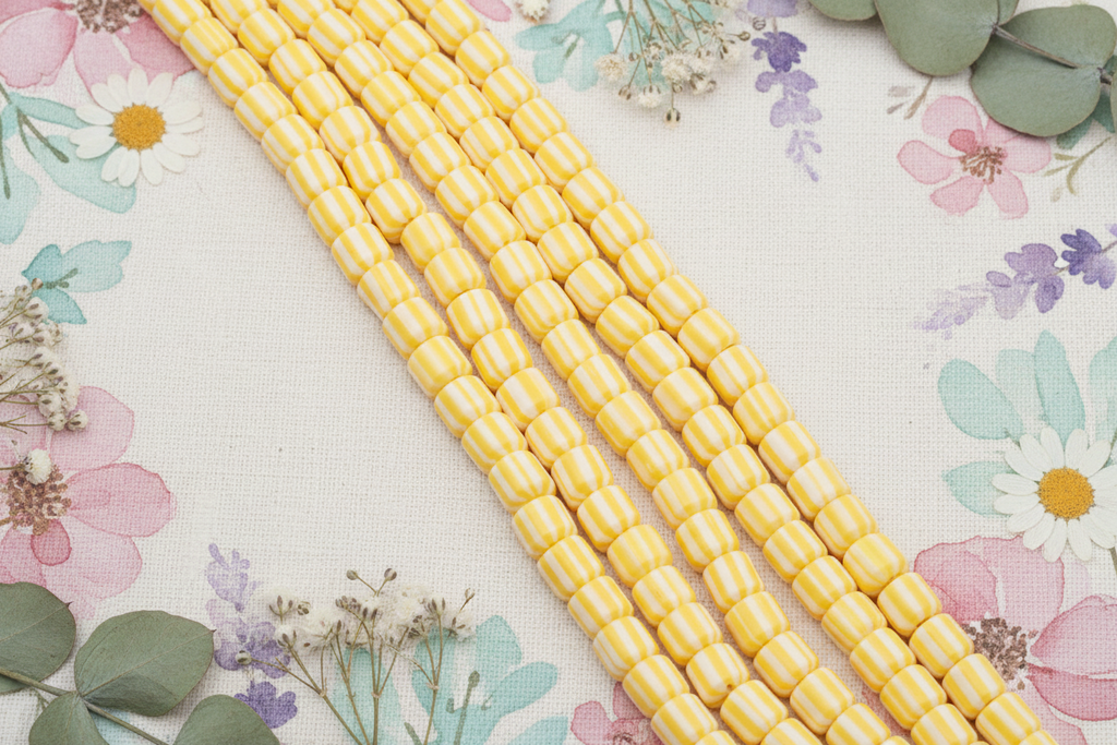 orange and white polymer clay barrel beads arranged diagonally on a floral fabric background with dried flowers and green leaves.