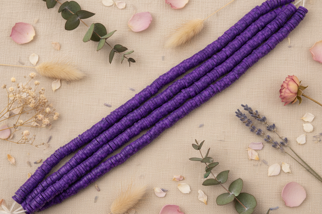 dakr purple speckled polymer clay disc beaded strands arranged diagonally on a beige fabric background, styled with dried flowers, greenery, and pink petals.