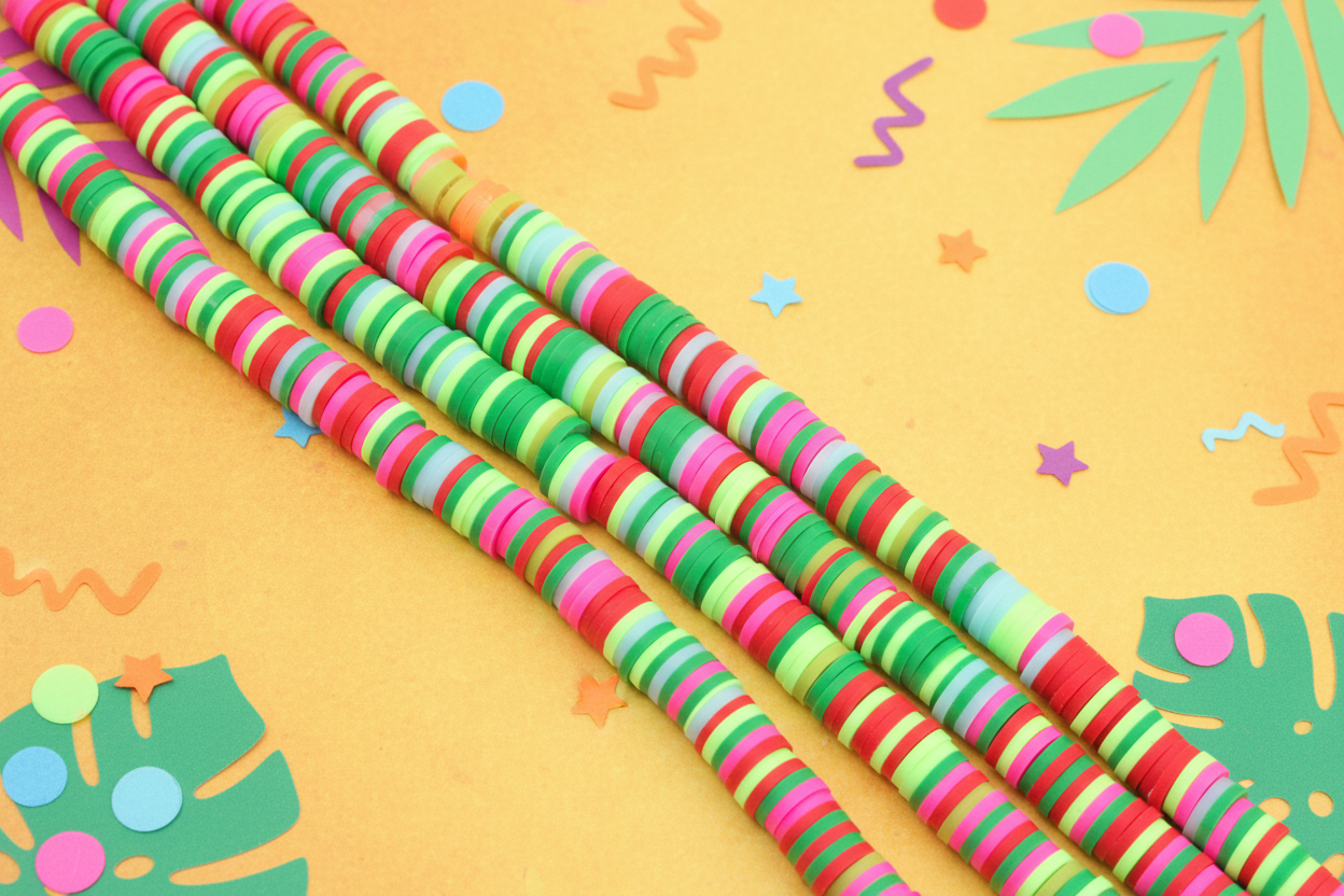 four strands of green and pink polymer clay disc beads, arranged diagonally on a yellow tropical-themed paper background.