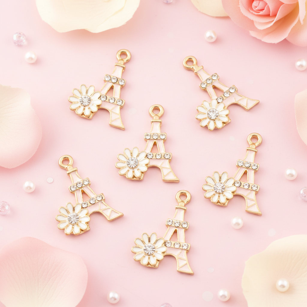 Five pink Eiffel Tower charms decorated with white enamel flowers and clear rhinestones, arranged on a pink background with soft rose petals and pearl-like beads.