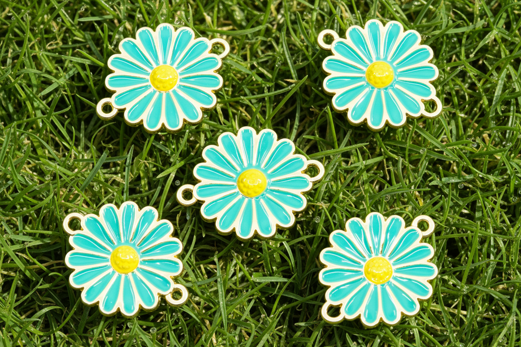 Five blue and yellow enamel flower charms with gold-tone metal edges and small hanging loops, arranged on green grass.