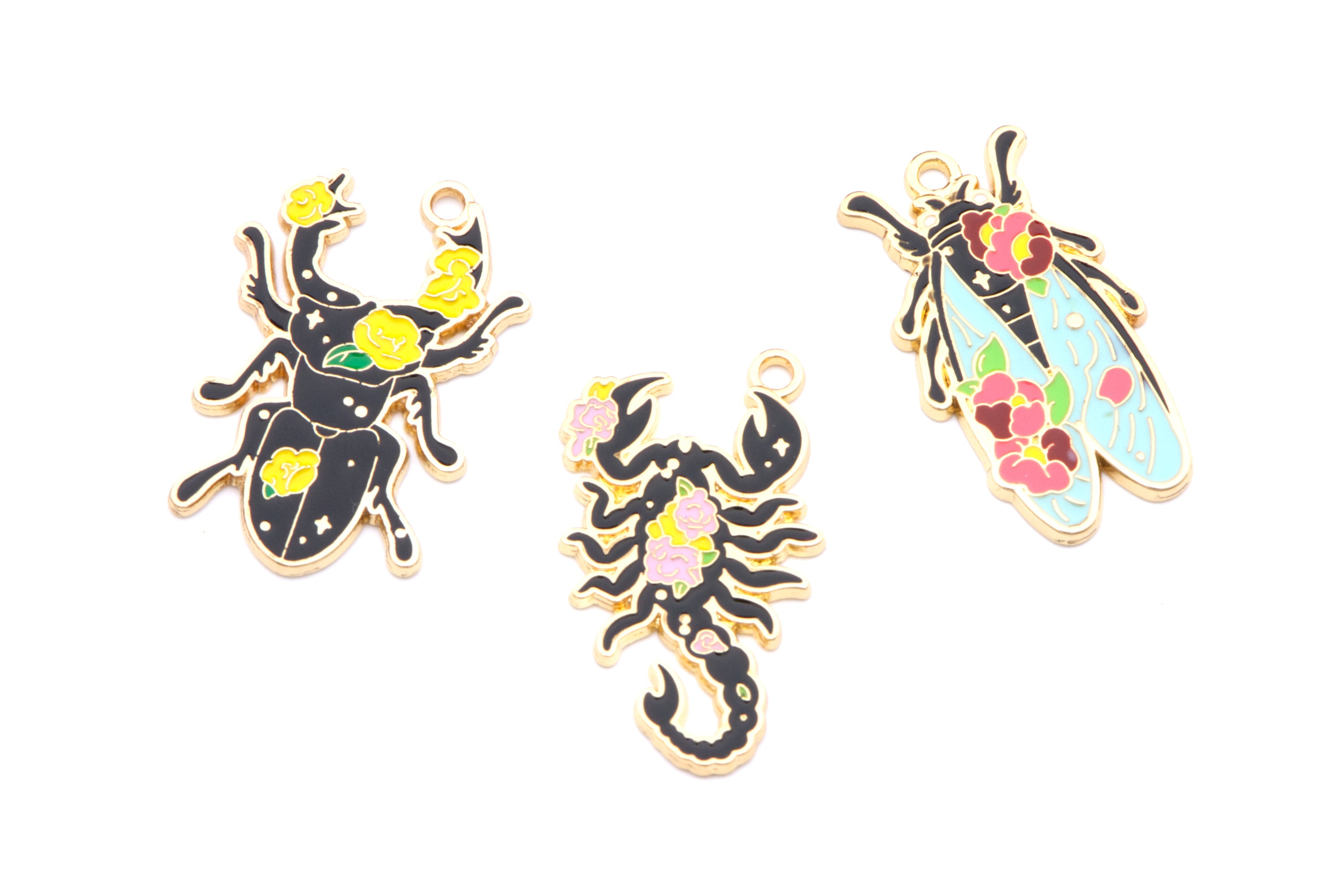 Three insect-shaped jewelry charms on a plain white background: a black beetle with yellow flowers, a black scorpion with pink flowers, and a light blue cicada with pink and red flowers.
