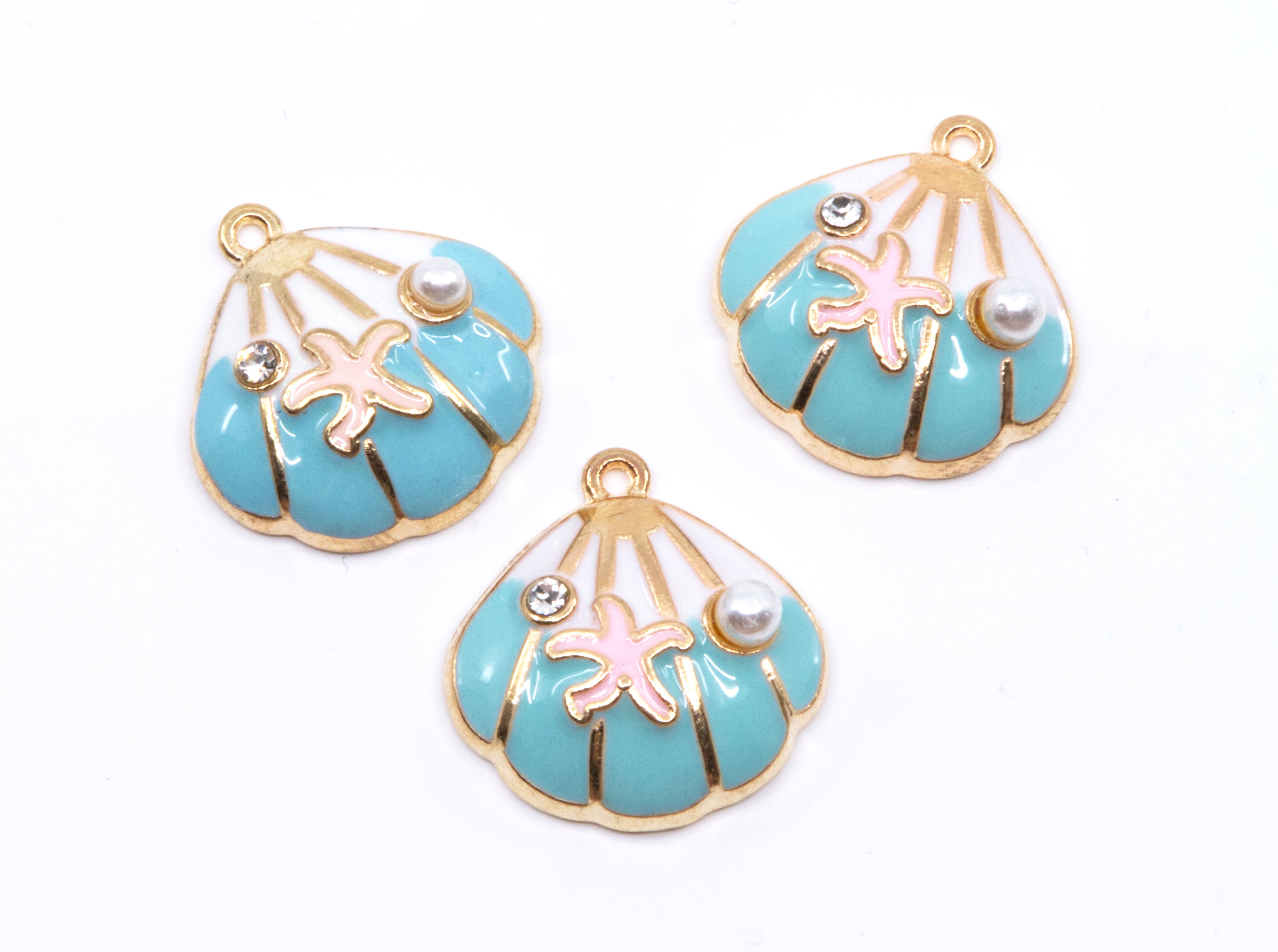 Three seashell-shaped jewelry charms with turquoise blue enamel, gold-tone metal borders, pink starfish details, and small pearl accents on a white background.