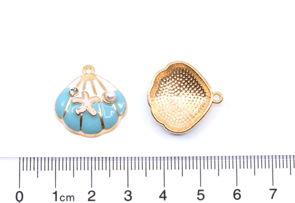 Two small blue and white seashell-shaped jewelry charms shown on a white background with a ruler at the bottom for scale. 
