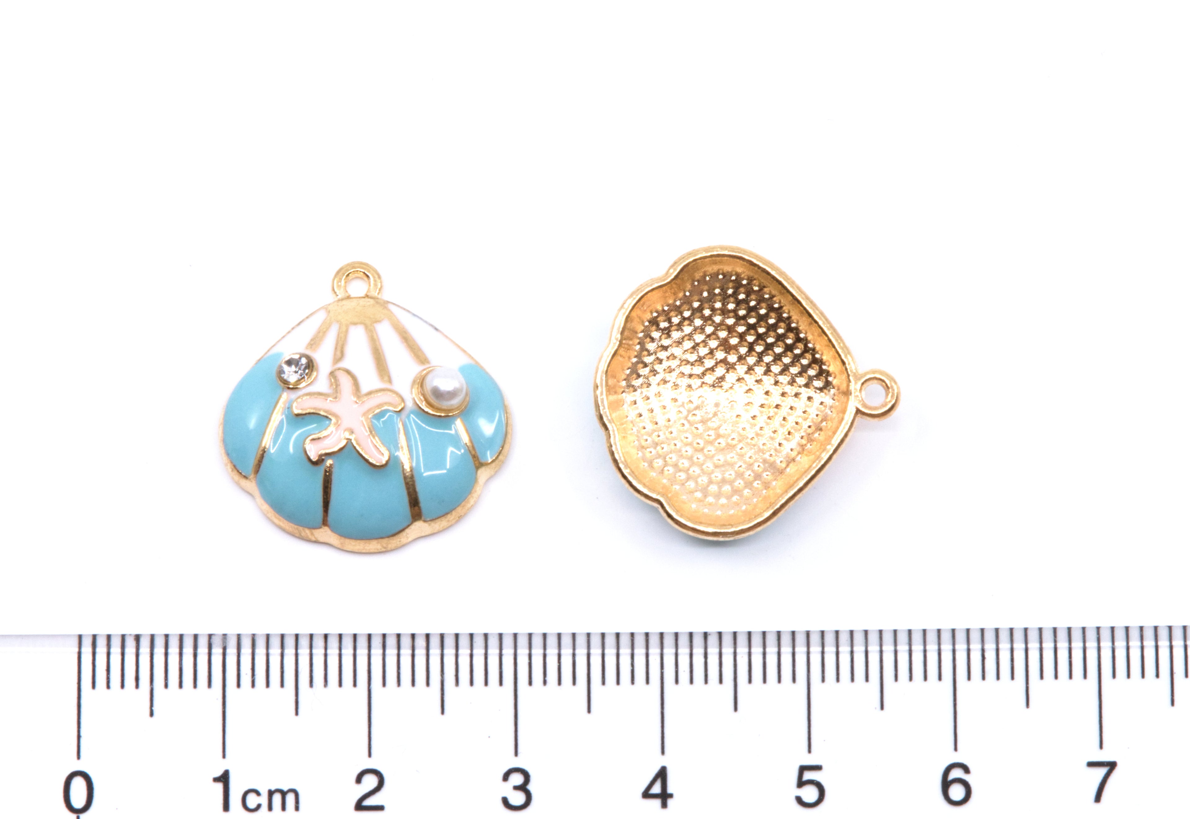 Two small blue and white seashell-shaped jewelry charms shown on a white background with a ruler at the bottom for scale. 