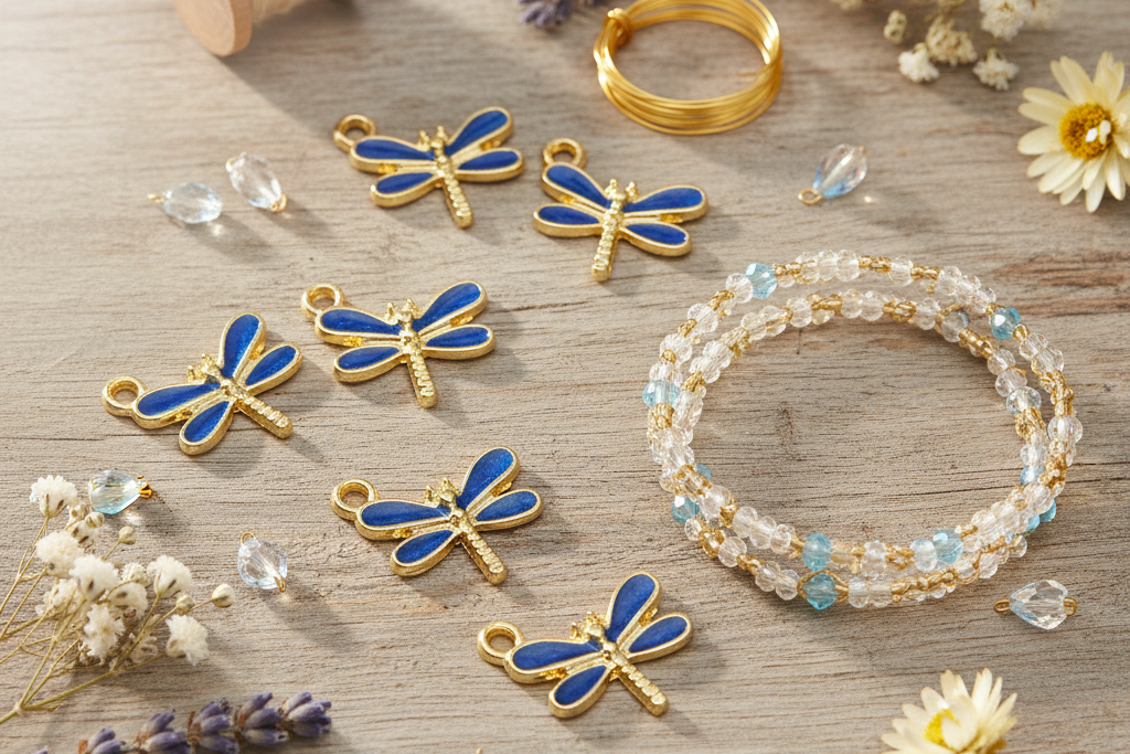 Dragonfly-shaped jewelry charms with blue enamel wings and gold-tone bodies, arranged on a light wood surface alongside a spool of white thread, gold wire, clear beads, and dried flowers.