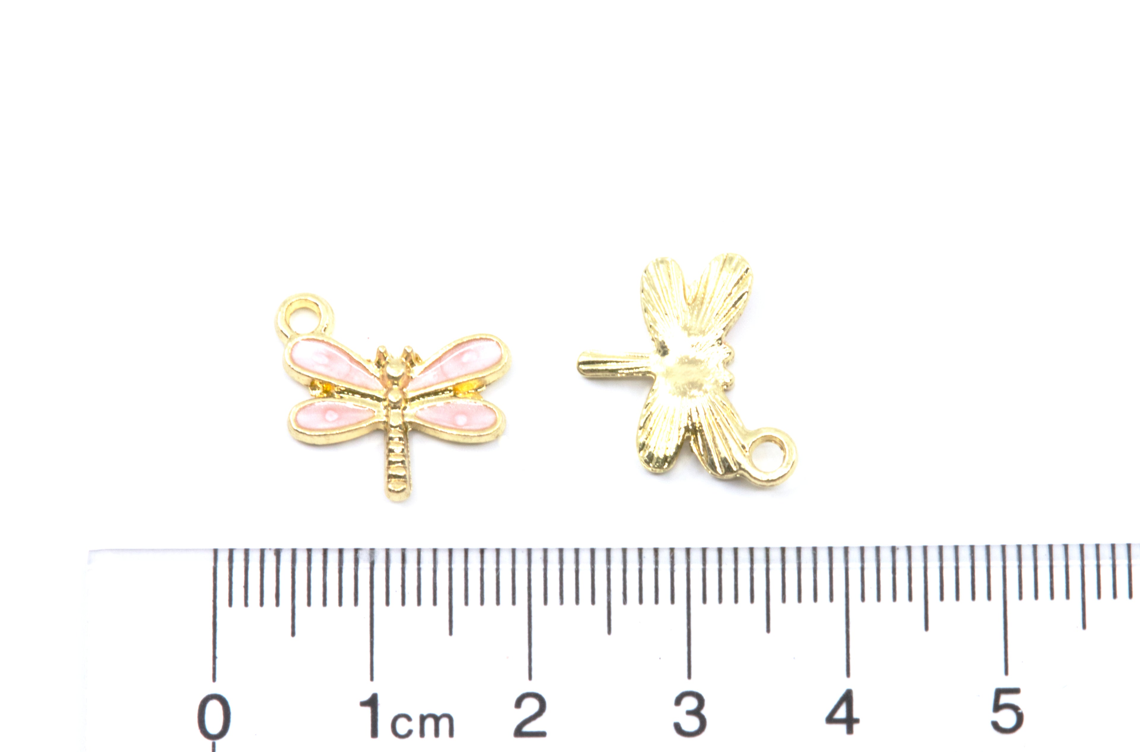 Two small dragonfly jewelry charms, one showing the front with pink enamel wings and the other showing the back with a loop, displayed above a cm ruler on a white background.