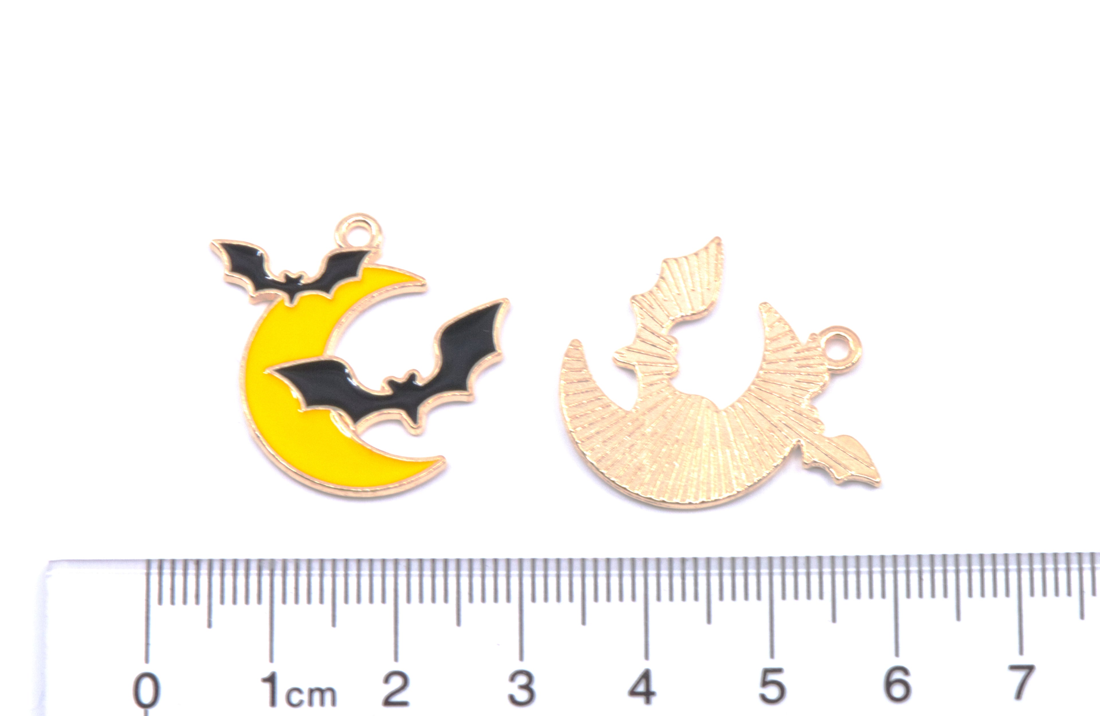 Two charms shaped like a crescent moon with a bat motif, shown on a white background above a ruler for scale. 