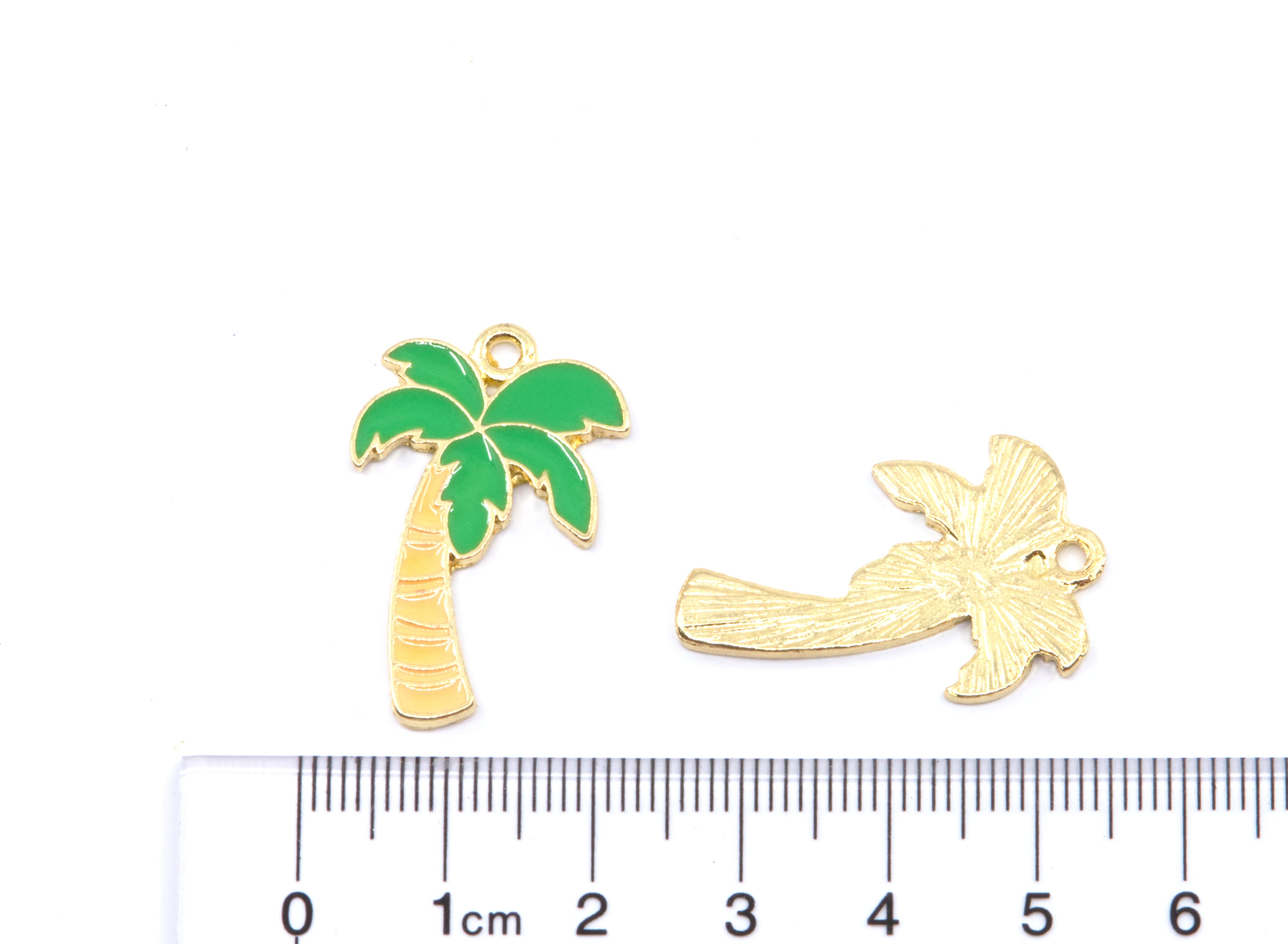 Two small gold-tone enamel jewelry charms, a palm tree with green leaves and a light brown trunk displayed above a cm ruler on a white background.