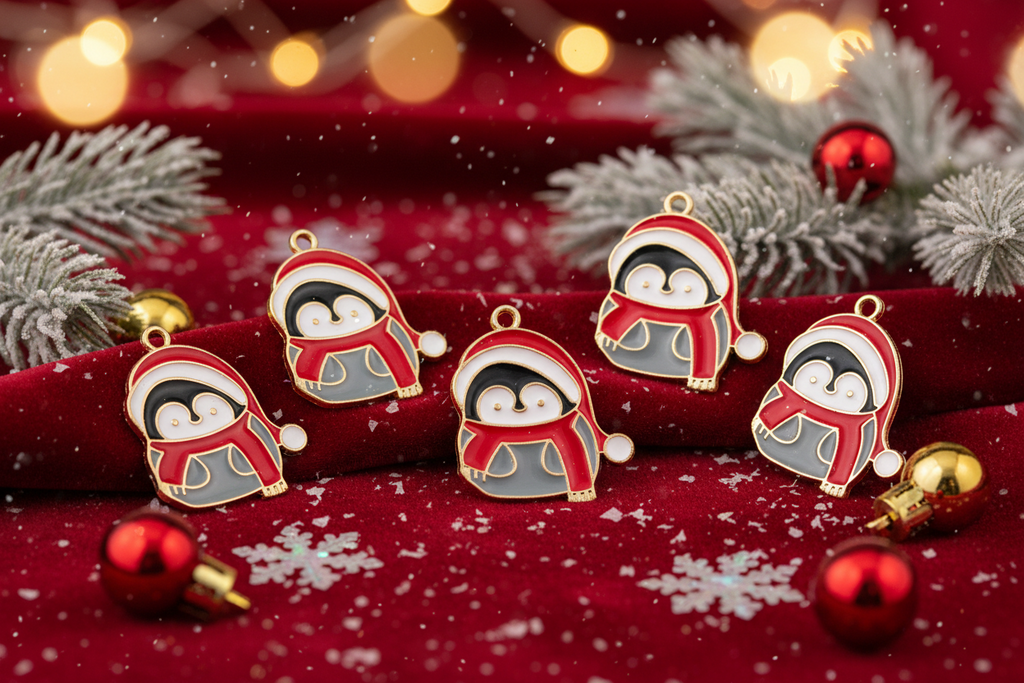 Five penguin-shaped Christmas charms with red Santa hats and scarves are arranged on a red fabric surface with holiday decorations, including silver pine branches, red ornaments, and warm bokeh lights in the background.