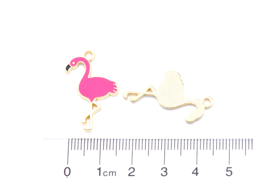 Two small flamingo-shaped jewelry charms on a white background, with one facing forward in bright pink and one facing backward in gold-tone metal. A ruler at the bottom shows the scale of the pieces.