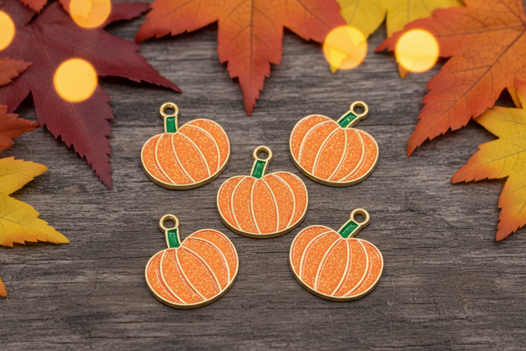 Five pumpkin-shaped charms with gold-tone metal borders and green stems, arranged on a dark wood surface with autumn leaves and warm bokeh lights around the edges.