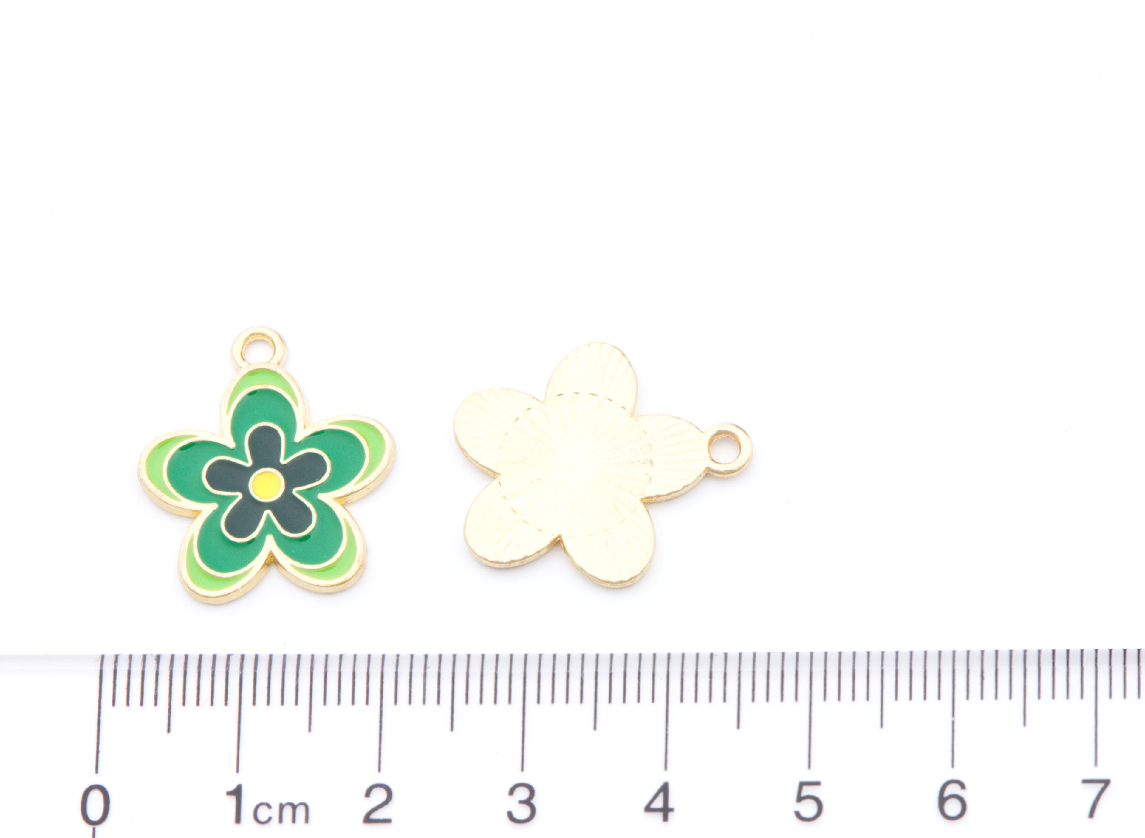 Two small flower-shaped jewelry charms shown on a white background with a ruler at the bottom for scale. One charm is front-facing and decorated with green, black, and yellow enamel, while the other is the plain back side in a pale gold tone.