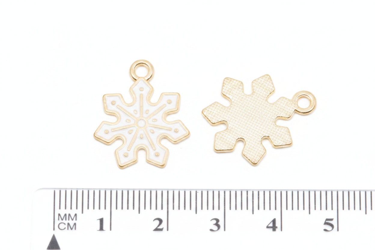 Two small snowflake-shaped jewelry charms with gold-tone edges and white enamel centers, shown next to a cm ruler on a white background.