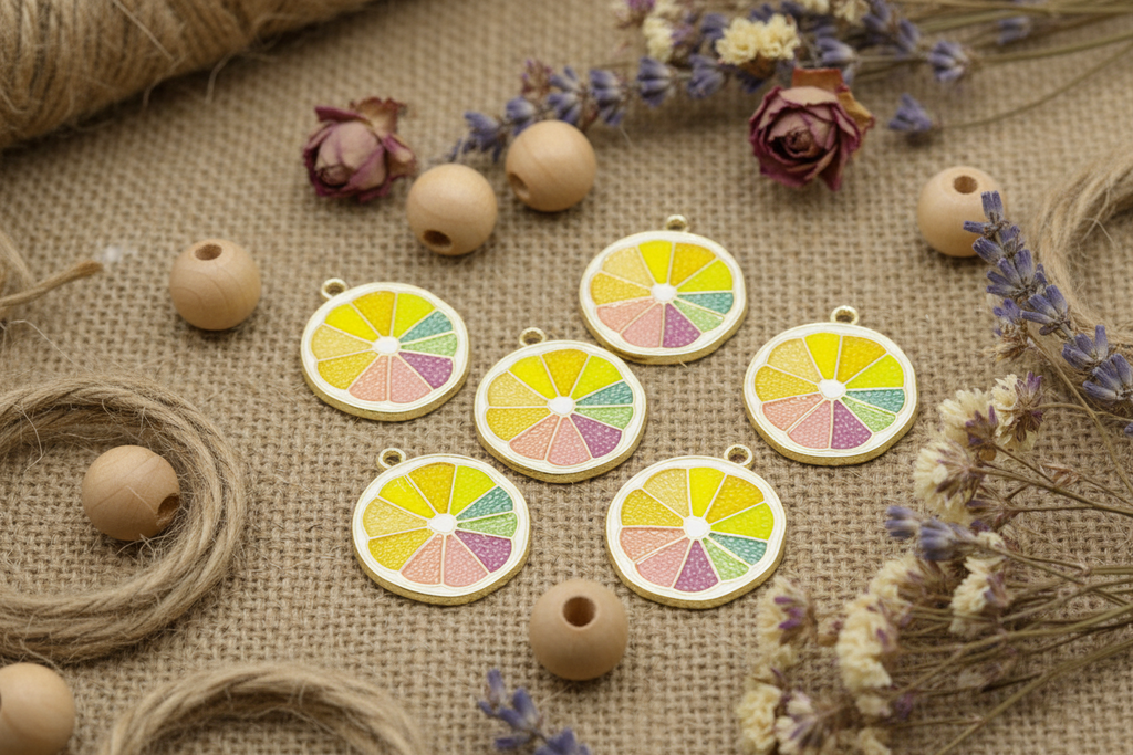 Six fruit slice enamel charms arranged on a burlap fabric background with wooden beads, twine, and dried flowers.