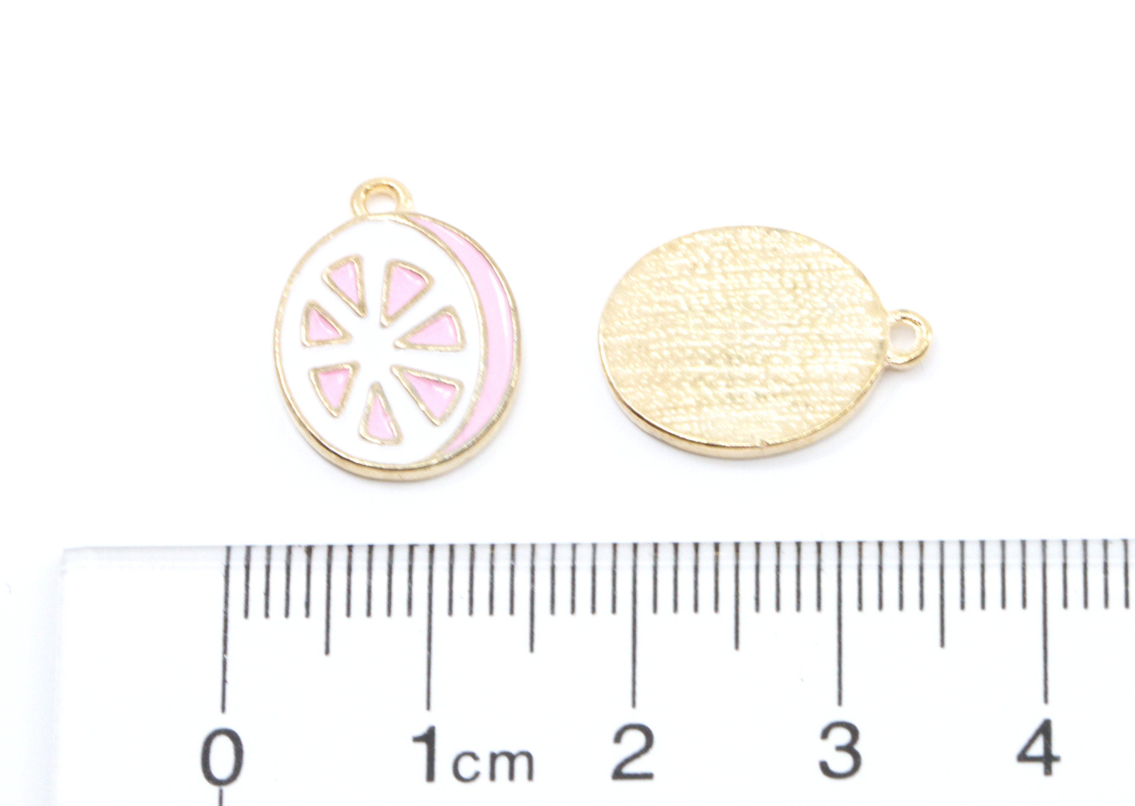 Two pink and white grapefruit slice jewelry charms shown on a white background with a ruler at the bottom for scale. 