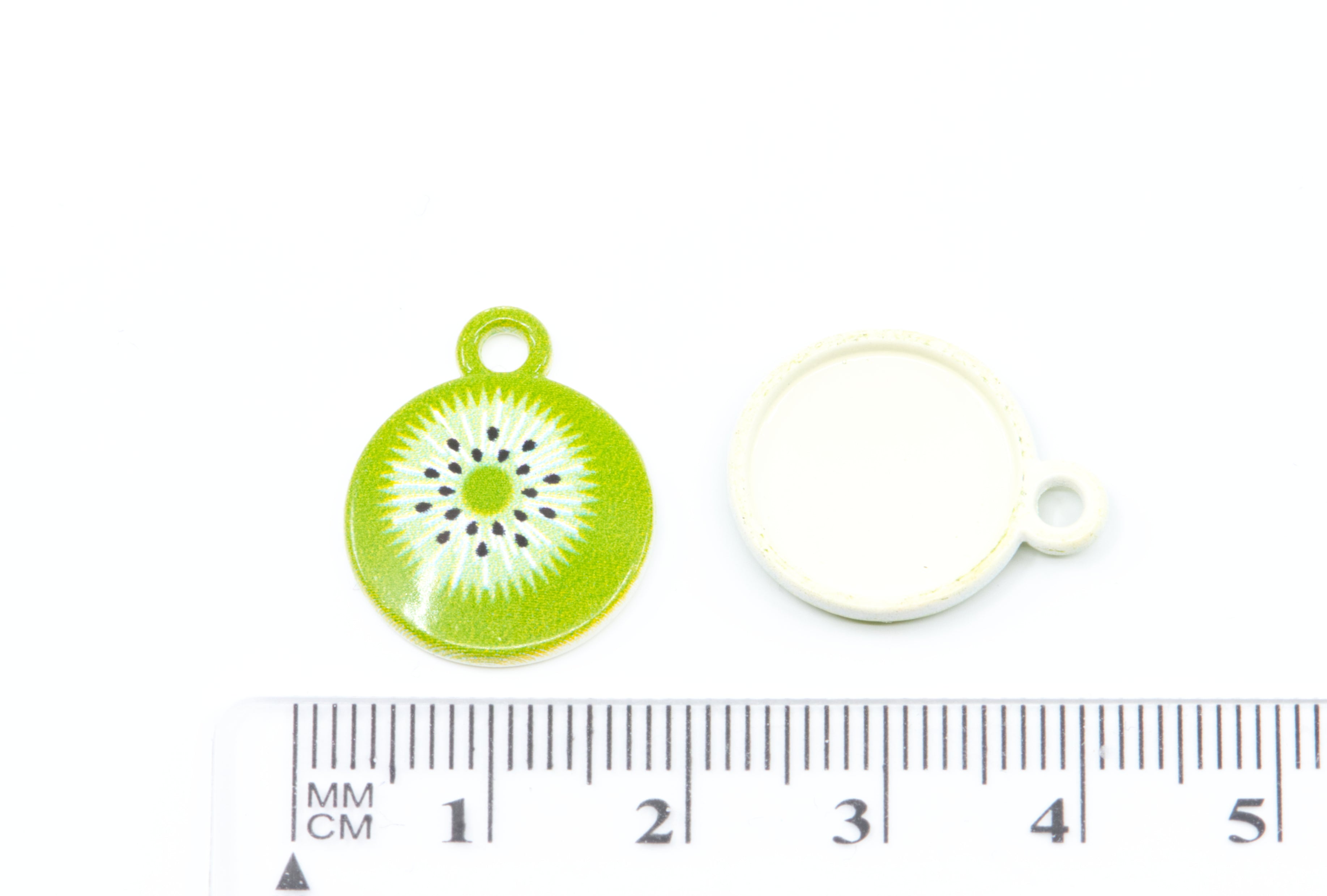 two kiwi slice charm with a green and white pattern and black seeds, shown next to a white backing disc and a ruler for scale on a white background.