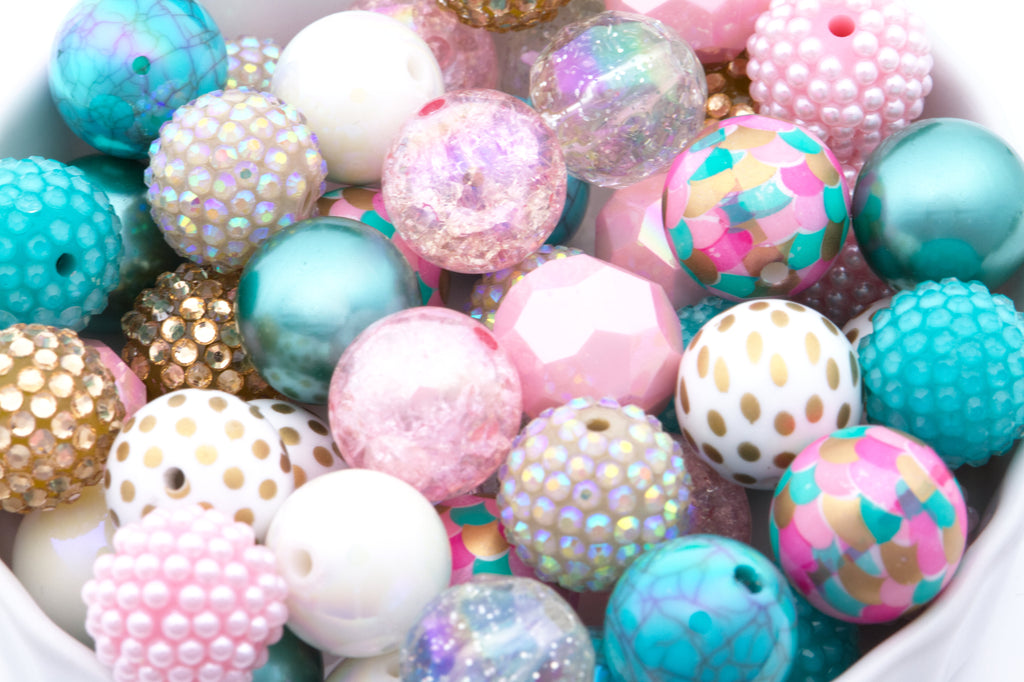 A close-up pile of colorful 20mm craft beads in various shapes and finishes, including pastel pink, turquoise, white, gold, and iridescent glitter, filling a white container.