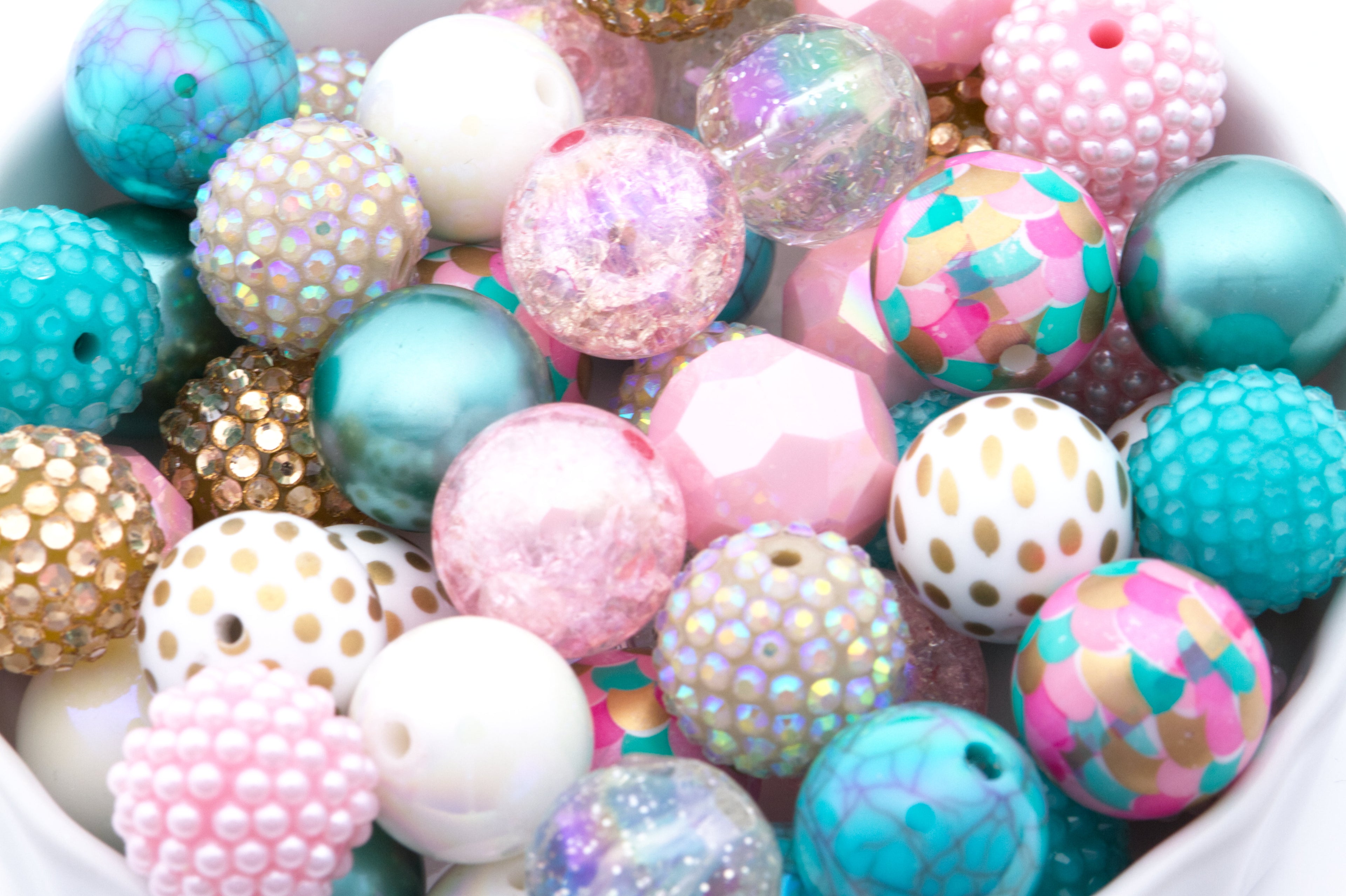 A close-up pile of colorful 20mm craft beads in various shapes and finishes, including pastel pink, turquoise, white, gold, and iridescent glitter, filling a white container.