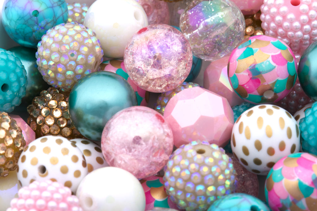 A close-up pile of colorful 20mm craft beads in various shapes and finishes, including pastel pink, turquoise, white, gold, and iridescent glitter, filling a white container.