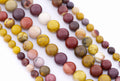 Multiple strands of polished round mookaite gemstone beads in assorted earthy colors including yellow, burgundy, cream, pink, and brown, arranged diagonally on a white background.