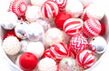 A close-up of assorted Christmas beads in a white bowl, featuring red, white, and silver holiday beads with striped, chevron, glitter, and textured finishes.