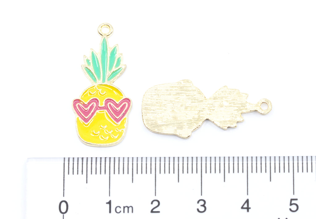 A pineapple-shaped jewelry charm with yellow enamel, green leaves, and pink heart-shaped sunglasses, shown next to its gold-toned back side and a ruler for scale on a white background.