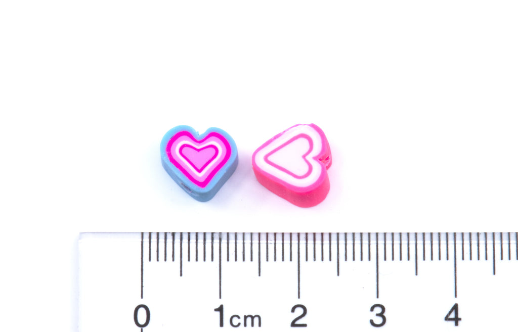 Two small heart-shaped polymer clay craft charms, one blue with pink and white concentric hearts and one pink with white and pink concentric hearts, shown next to a cm ruler for scale on a white background.