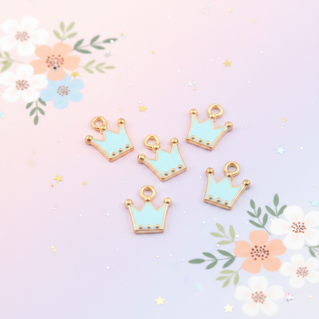 Five small crown-shaped jewelry charms with light blue enamel and gold-tone edges, arranged on a pastel pink background with floral and star confetti decorations.