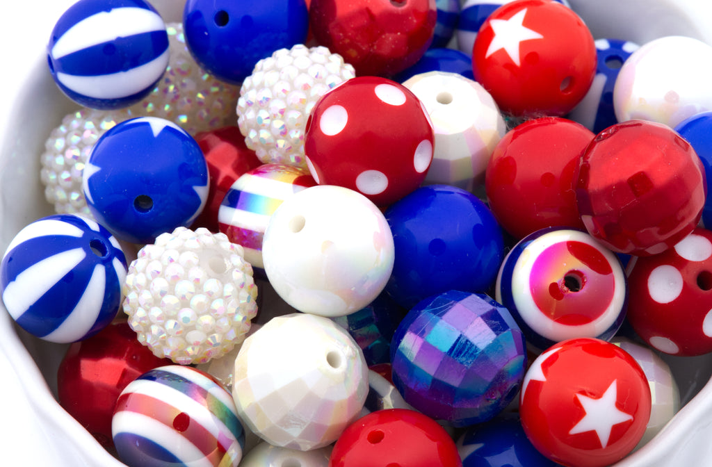 A close-up of assorted decorative beads in red, white, and blue, including striped, polka-dot, faceted, and iridescent styles, filling a white container.