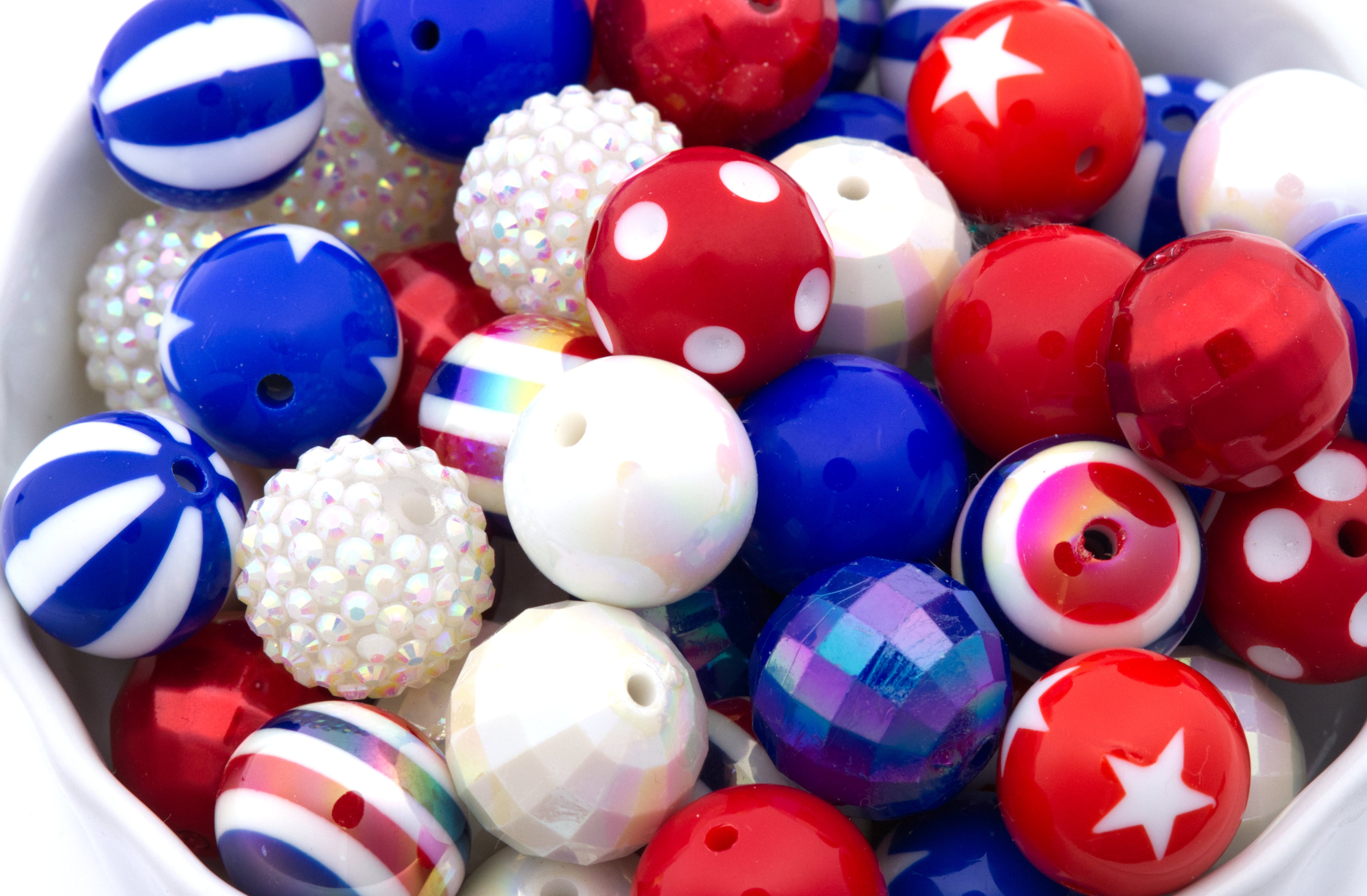 A close-up of assorted decorative beads in red, white, and blue, including striped, polka-dot, faceted, and iridescent styles, filling a white container.