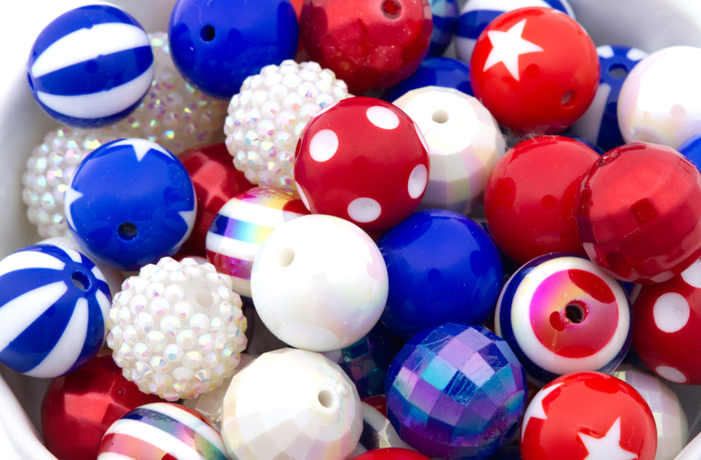 A close-up of assorted decorative beads in red, white, and blue, including striped, polka-dot, faceted, and iridescent styles, filling a white container.