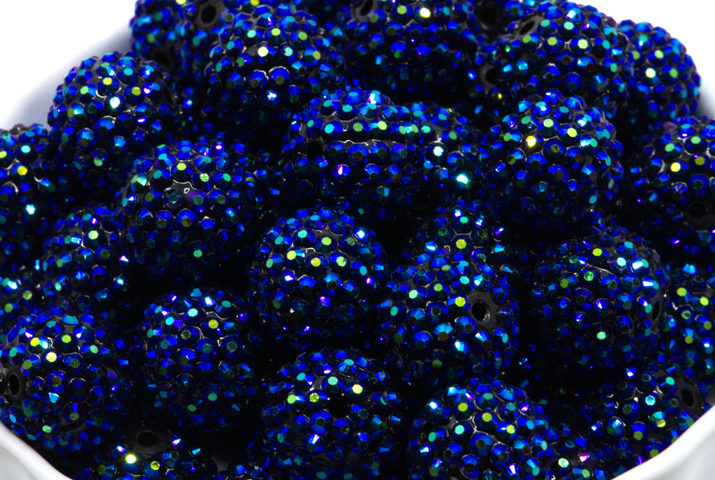 A close-up of multiple metallic blue rhinestone covered beads piled in a white container.