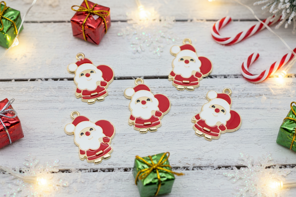 Five Christmas-themed enamel charms shaped like Santa Claus are arranged on a white wooden surface with festive props including wrapped gift boxes, candy canes, and warm fairy lights.