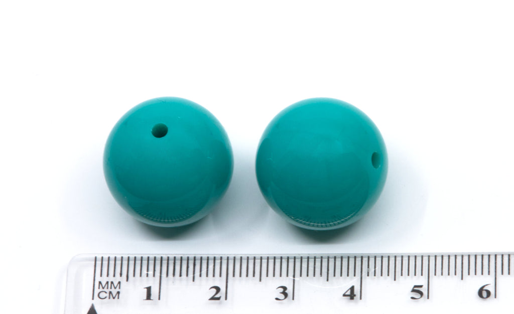 Two teal beads next to a ruler for scale on a white background