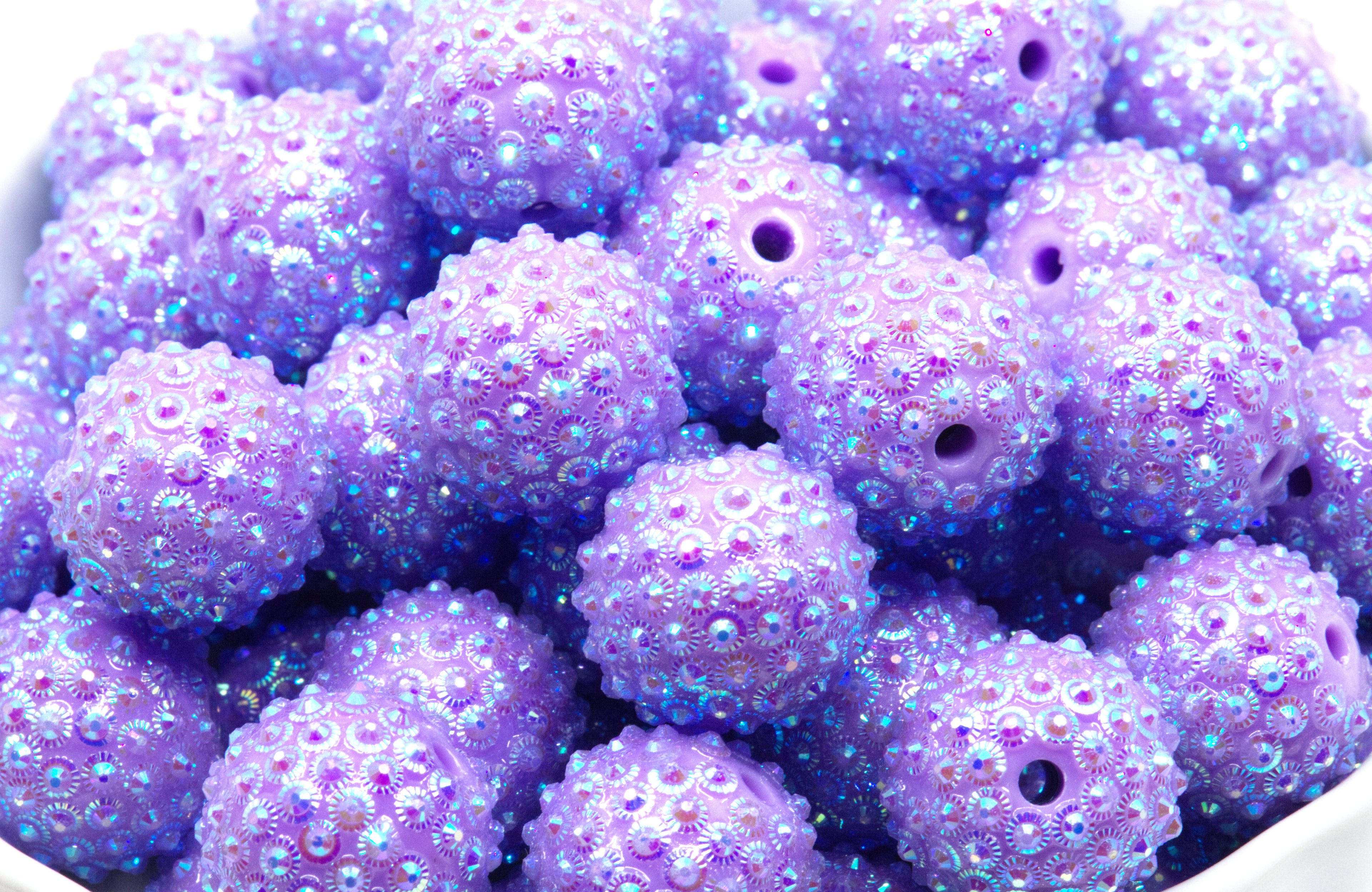 A close-up pile of purple sunburst rhinestone beads in a white bowl on a white background. 