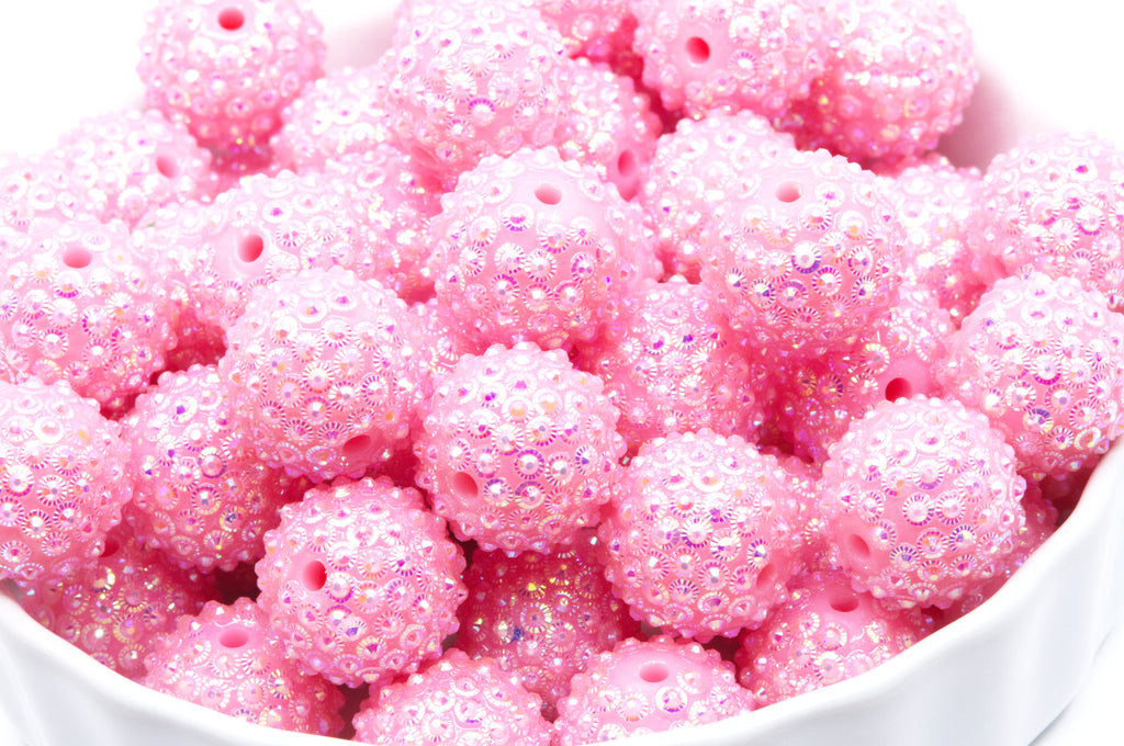 A close-up of pink rhinestone bubblegum beads, piled in a white bowl against a white background.