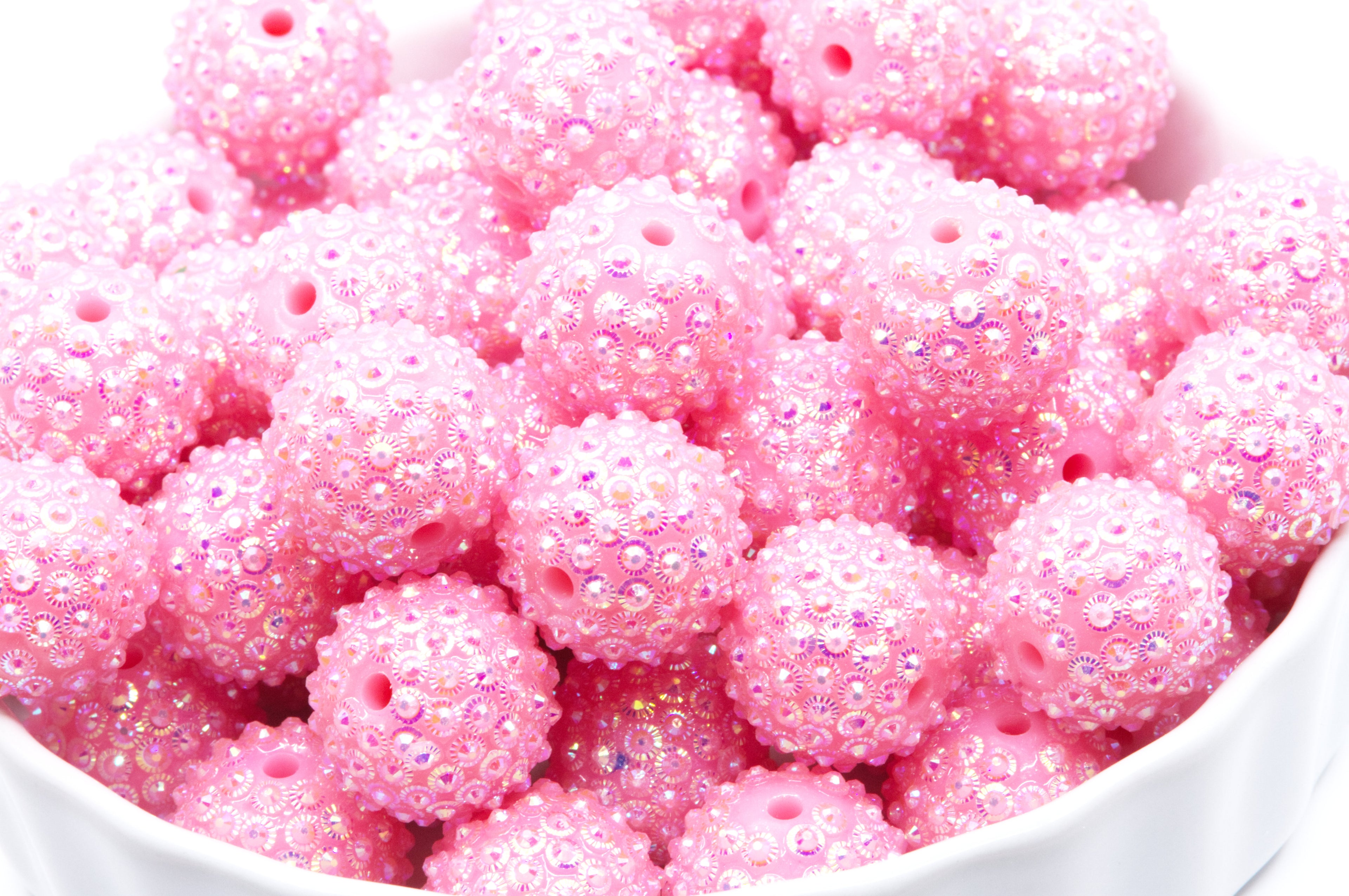A close-up of pink rhinestone bubblegum beads, piled in a white bowl against a white background.