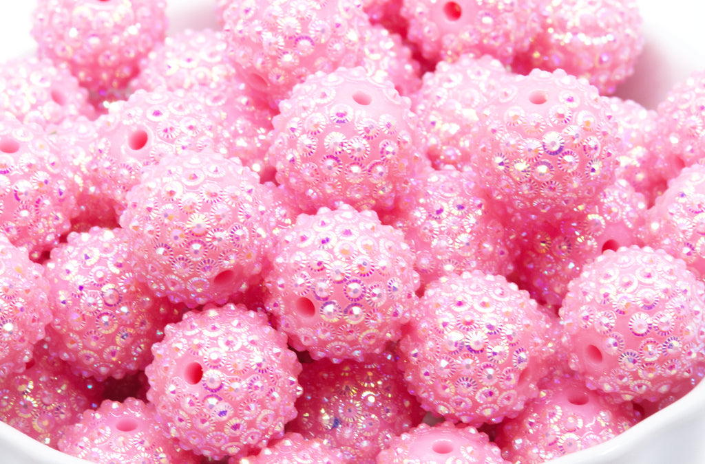 A close-up of pink rhinestone bubblegum beads, piled in a white bowl against a white background.