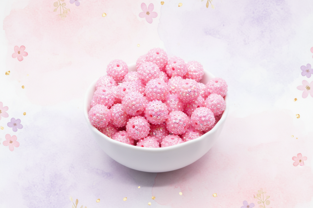 A white ceramic bowl filled with pink rhinestone bubblegum beads, set against a pastel pink and purple watercolor background decorated with small floral motifs.
