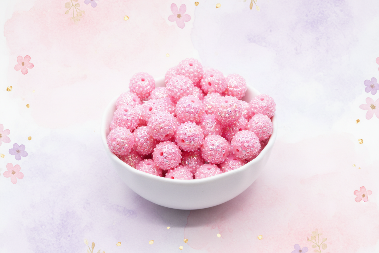 A white ceramic bowl filled with pink rhinestone bubblegum beads, set against a pastel pink and purple watercolor background decorated with small floral motifs.