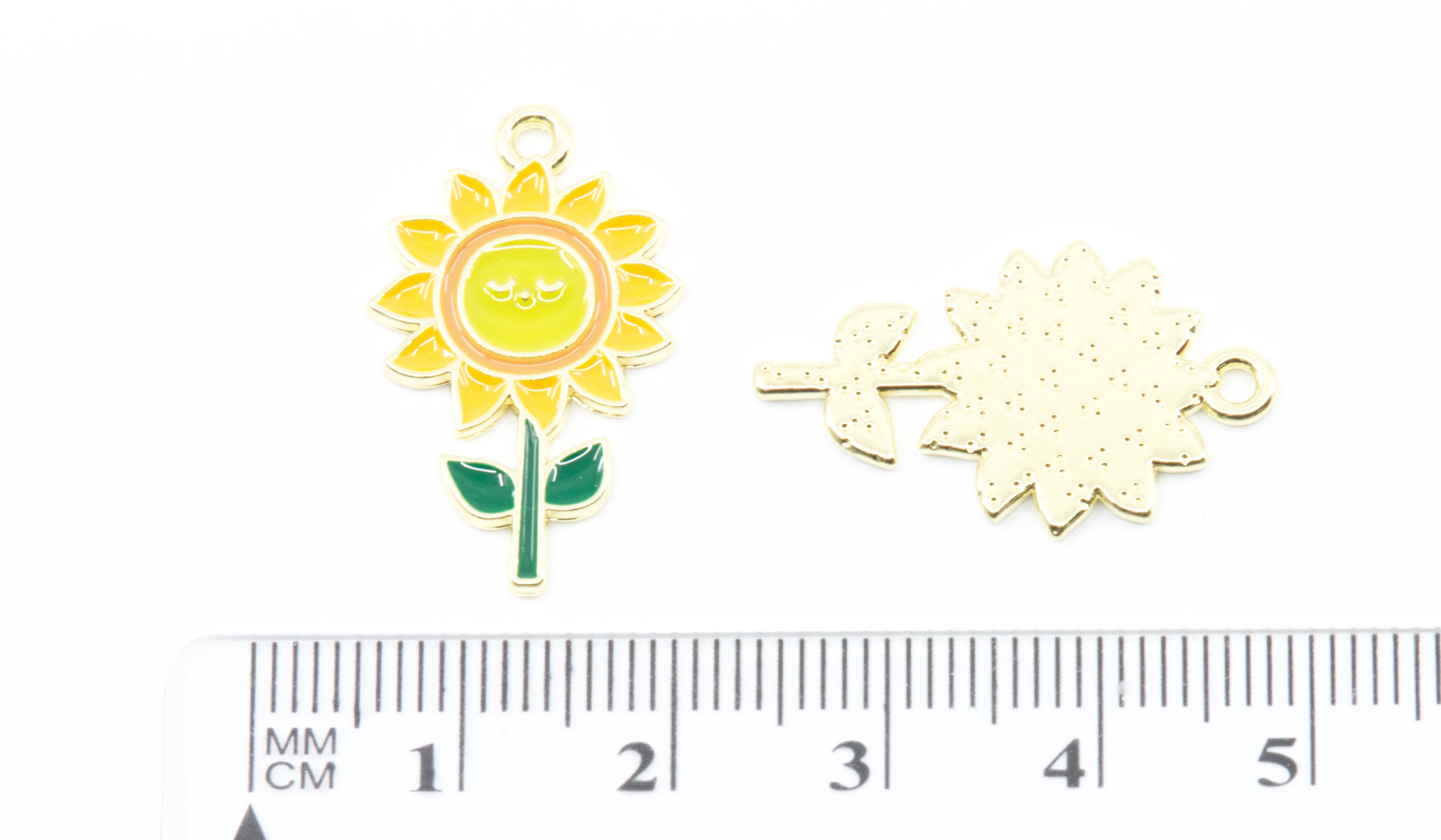 Two small enamel flower charms on a white background, with a cm ruler below for scale. One charm shows a yellow sunflower with green leaves and a smiling face, and the other shows the reverse side with a textured gold-toned backing.