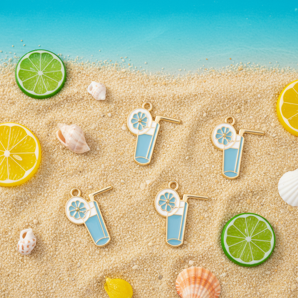 Four cocktail-themed enamel charms with gold-tone metal borders are arranged on a sandy beach background with tropical props including sliced limes, a lemon wedge, and seashells. The background features a gradient from beige sand to a blue ocean water scene.