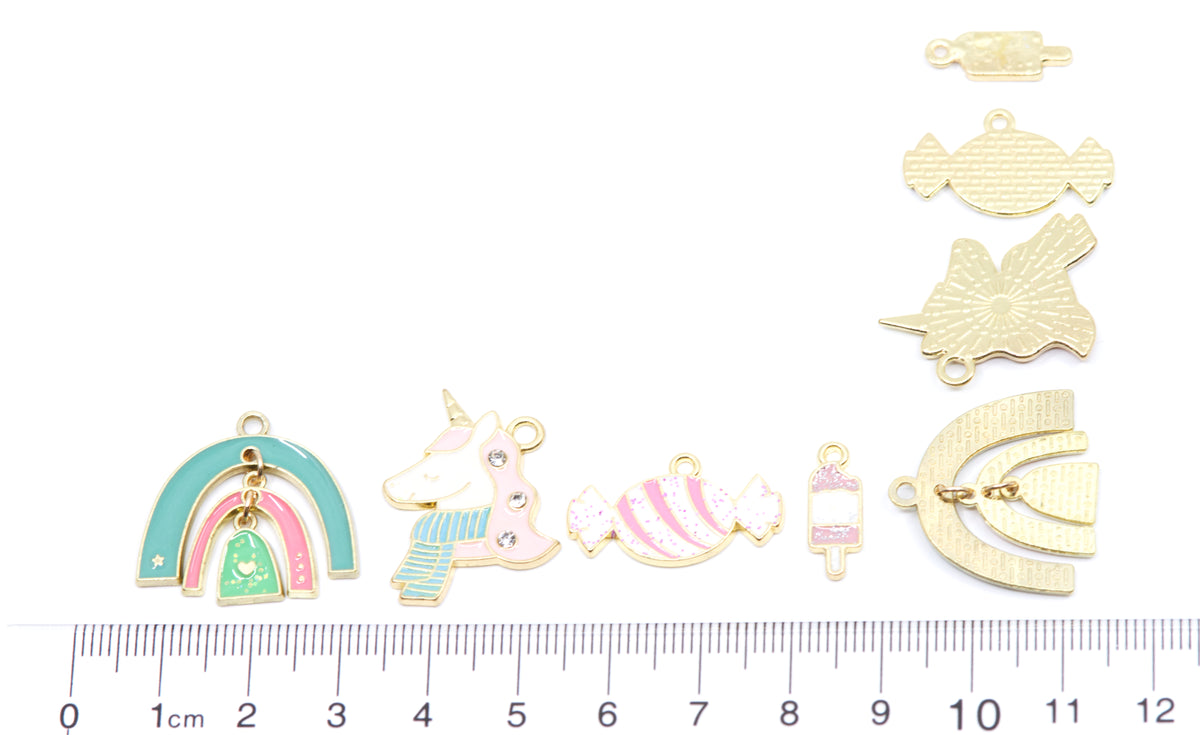 A set of small enamel jewelry charms and findings laid out on a white background with a  cm ruler at the bottom for scale. The charms include a pastel rainbow, a unicorn, pink-and-white candy shapes, a small pink popsicle, 